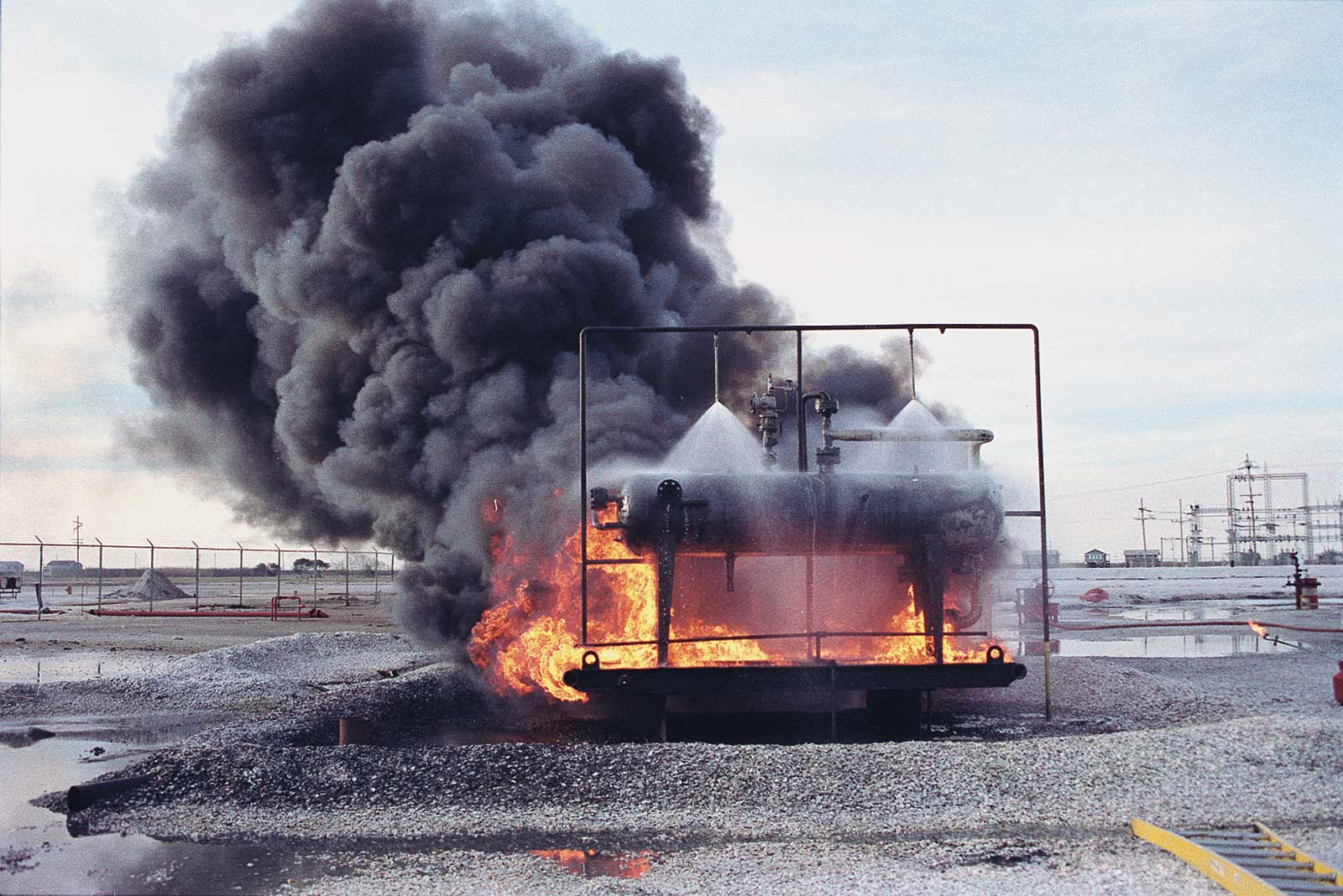 Chemical tank fire protection test of two spiral fire protection nozzles mounted on water pipes spraying a deluge of water onto a small chemical tank surrounded by flames